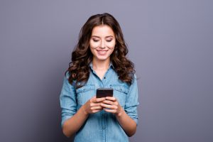 Content trendy cheerful beautiful beautiful charming adorable attractive brunette girl with wavy hair in casual denim shirt, typing on phone, isolated over gray background