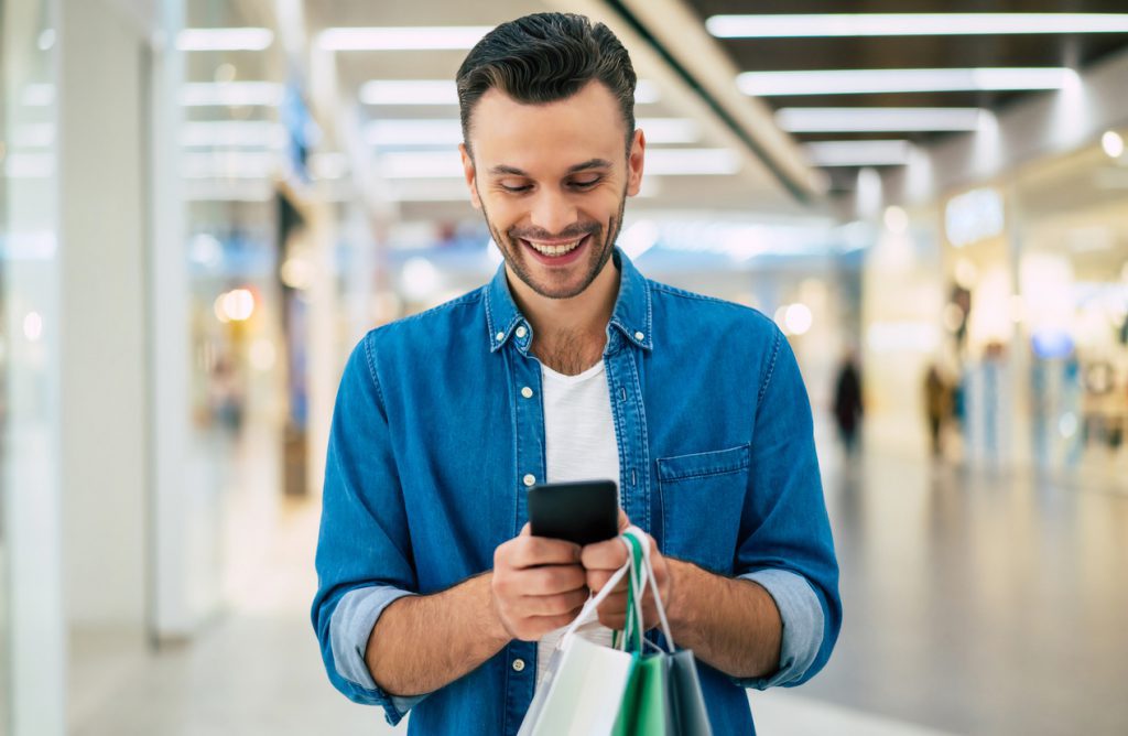 مرد ریشوی جوان و خوش‌تیپ خندان با کیف‌های خرید با استفاده از تلفن هوشمند در حالی که در جمعه سیاه در مرکز خرید قدم می‌زند